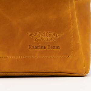 Puede incluir: Primer plano de un bolso de cuero marr&oacute;n con el texto "MG Leather Work" y "Kaarina Beam" grabado en relieve en la parte delantera.