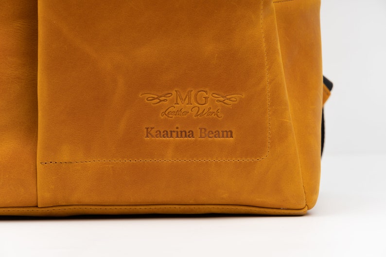Puede incluir: Primer plano de un bolso de cuero marr&oacute;n con el texto "MG Leather Work" y "Kaarina Beam" grabado en relieve en la parte delantera.