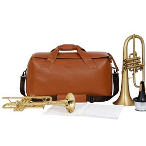 May include: A brown leather trumpet case with a black strap and two gold trumpets. The case has a zipper on the top and a handle on the side.