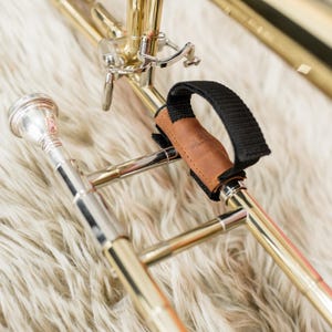 May include: A brass trombone slide featuring a silver mouthpiece and a brown leather hand grip with a black nylon strap attached to the metal tubing. The instrument displays polished metallic surfaces and mechanical components including a water key.
