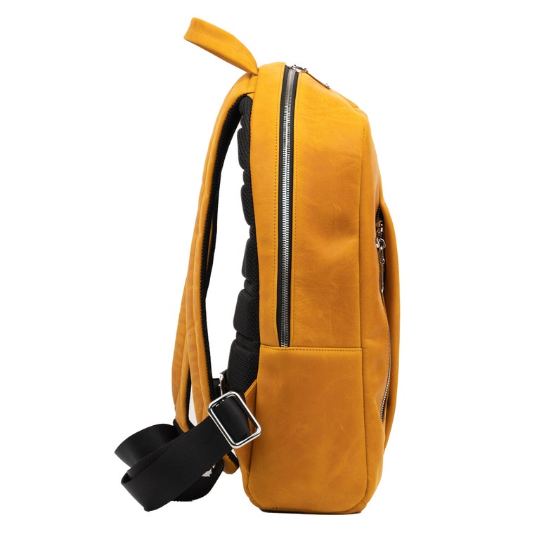 Puede incluir: Una mochila de cuero amarillo mostaza con correas negras y una cremallera plateada. La mochila tiene un panel trasero acolchado y un peque&ntilde;o bolsillo con cremallera en la parte delantera.