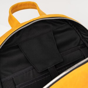 Puede incluir: Una mochila de cuero amarilla con una abertura con cremallera. El interior de la mochila est&aacute; forrado con tela negra y tiene varios bolsillos.