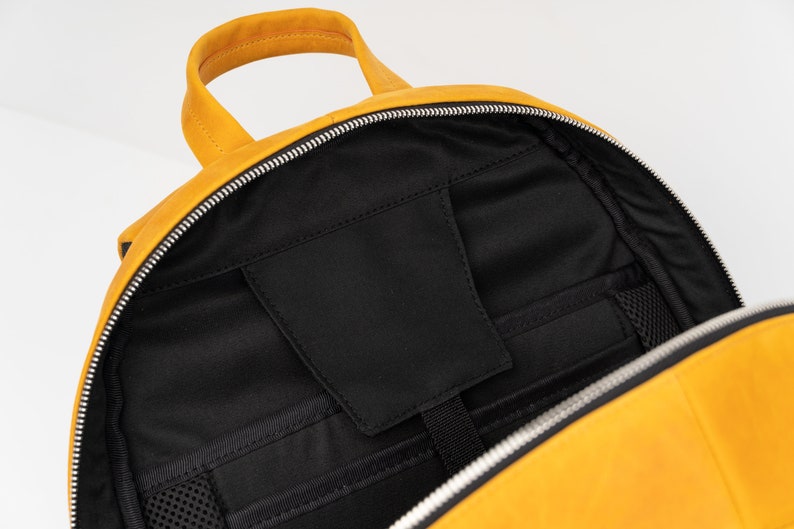 Puede incluir: Una mochila de cuero amarilla con una abertura con cremallera. El interior de la mochila est&aacute; forrado con tela negra y tiene varios bolsillos.