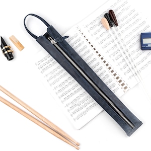 May include: An assortment of musical instruments and accessories, including a dark blue leather case with a zipper, a clarinet mouthpiece, drumsticks, and a digital tuner, all arranged on a sheet music background.