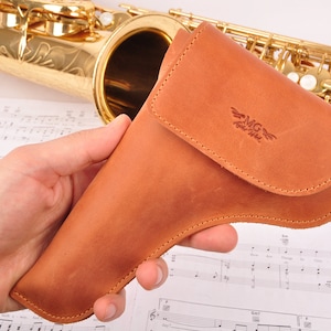 May include: A brown leather saxophone case with a stitched flap and the text "MG Leather" embossed on the front.