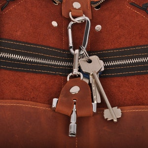 Peut inclure: Gros plan sur un sac en cuir marron avec une fermeture éclair noire et un mousqueton argenté tenant des clés. Le sac a une surface texturée et un petit porte-clés en cuir.