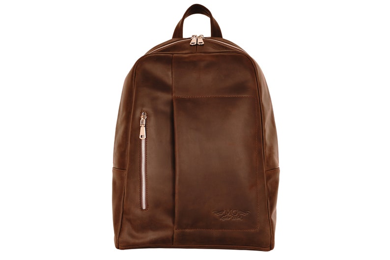 Puede incluir: Una mochila de cuero marr&oacute;n con un bolsillo con cremallera en la parte delantera y un peque&ntilde;o logotipo en la parte inferior.