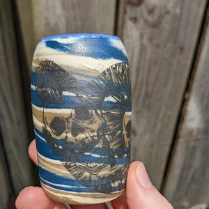 May include: A small, ceramic cup with a blue, white, and brown striped design. The cup features a black and white illustration of two skulls.