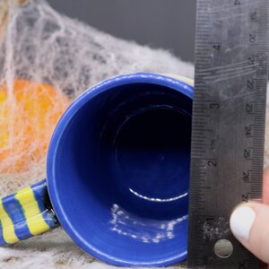 May include: A blue ceramic mug with a yellow and blue striped handle. The mug is 3 inches in diameter and is sitting on a white surface.