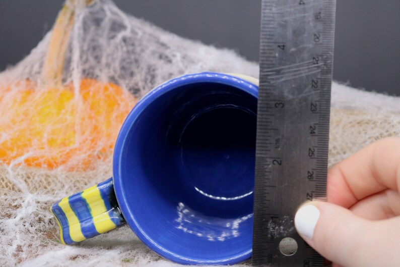 May include: A blue ceramic mug with a yellow and blue striped handle. The mug is 3 inches in diameter and is sitting on a white surface.