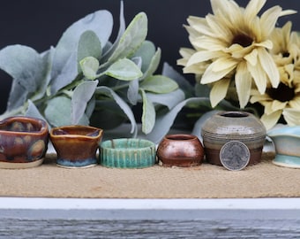 May include: A collection of six small, handmade ceramic pots in various colours and styles. The pots are arranged in a row on a brown surface. A UK 20 pence coin is placed in front of one of the pots for scale.