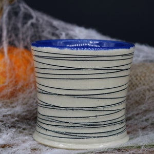 May include: A white ceramic mug with a blue rim and black lines. The mug is sitting on a white surface.