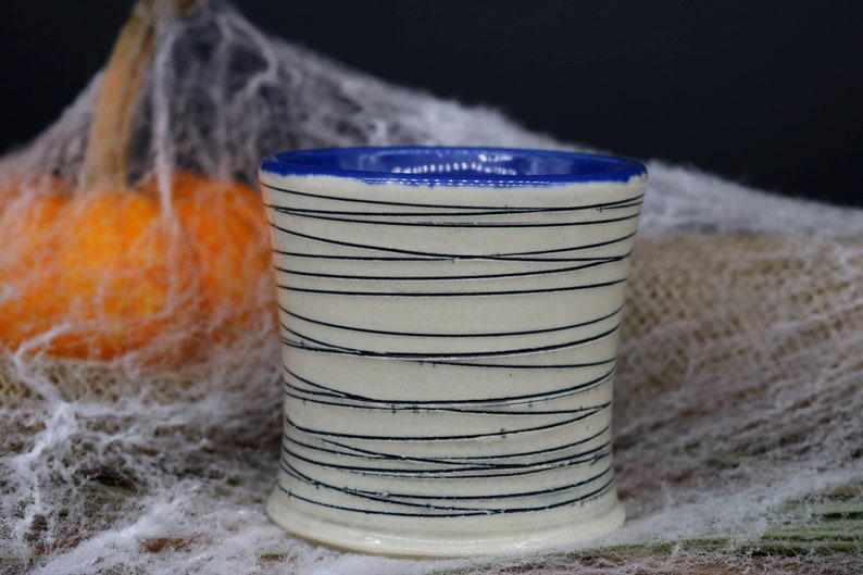 May include: A white ceramic mug with a blue rim and black lines. The mug is sitting on a white surface.