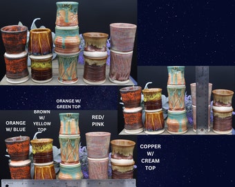 May include: A collection of handmade ceramic mugs in various colours and patterns, including orange with green top, brown with yellow top, orange with blue top, red/pink, and copper with cream top. The mugs are stacked in groups of three, with a ruler in the background for size reference.