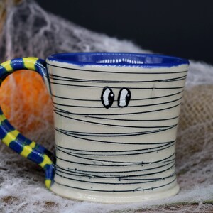 May include: A white ceramic mug with a blue rim and a yellow and blue striped handle. The mug is decorated with black lines to resemble a mummy and has two googly eyes.