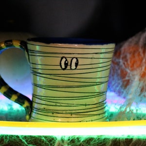 May include: A green and yellow striped ceramic mug with black eyes and a blue rim. The mug is sitting on a green and yellow surface with a white and orange pumpkin in the background.
