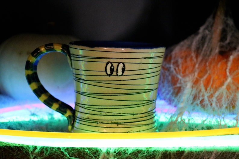 May include: A green and yellow striped ceramic mug with black eyes and a blue rim. The mug is sitting on a green and yellow surface with a white and orange pumpkin in the background.