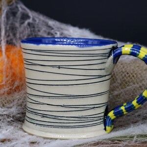 May include: A white ceramic mug with a blue rim and a yellow and blue striped handle. The mug has black lines drawn on the side.