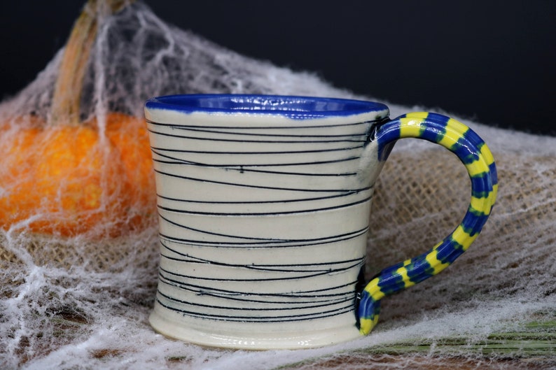 May include: A white ceramic mug with a blue rim and a yellow and blue striped handle. The mug has black lines drawn on the side.