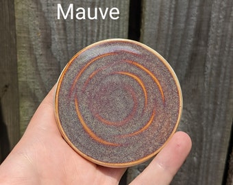 May include: A small, round ceramic coaster with a mauve glaze and concentric circles in a darker brown glaze.