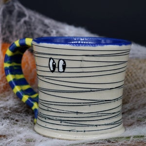 May include: A white ceramic mug with a blue rim and a yellow and blue striped handle. The mug is decorated with black lines to resemble a mummy and has two googly eyes.