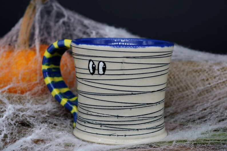May include: A white ceramic mug with a blue rim and a yellow and blue striped handle. The mug is decorated with black lines to resemble a mummy and has two googly eyes.