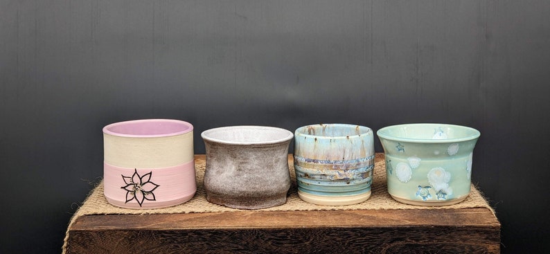 May include: Four ceramic mugs with different designs and colours. The mugs are arranged in a row on a wooden surface. The mugs are all different sizes and shapes. The mugs are all handmade and unique.