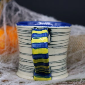 May include: A white ceramic mug with blue and yellow stripes on the handle. The mug has a white background with black stripes.