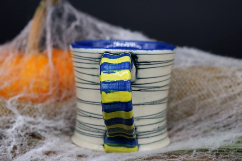 May include: A white ceramic mug with blue and yellow stripes on the handle. The mug has a white background with black stripes.