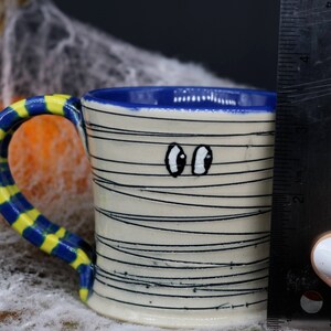 May include: A white ceramic mug with a blue rim and a yellow and blue striped handle. The mug is decorated with black lines to resemble a mummy and has two googly eyes painted on the front.