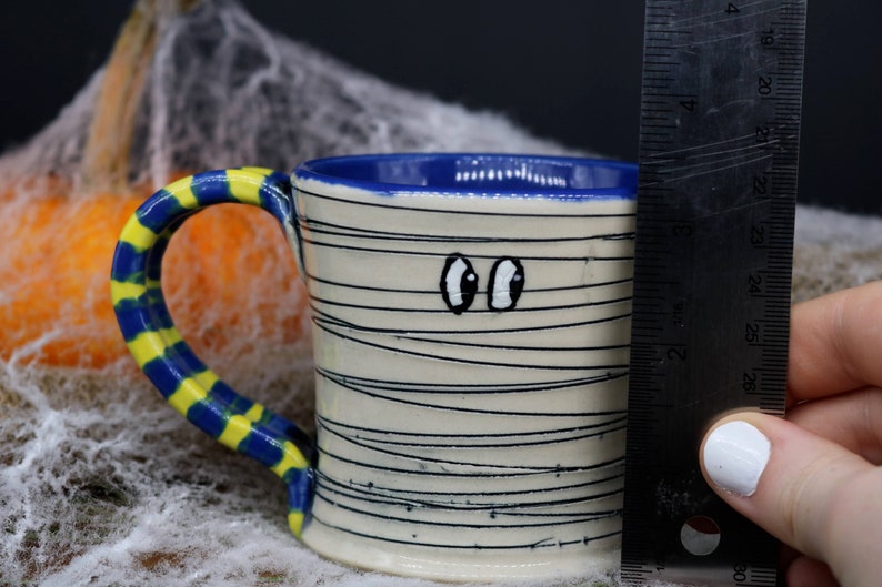 May include: A white ceramic mug with a blue rim and a yellow and blue striped handle. The mug is decorated with black lines to resemble a mummy and has two googly eyes painted on the front.