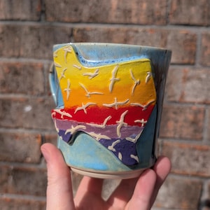 May include: A ceramic mug with a colorful design of a sunset with birds flying over it. The mug is hand-painted with a rainbow of colors, including yellow, orange, red, purple, and blue.