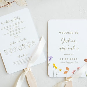 Op de afbeelding: Twee witte bruiloftsvaandels met gouden letters. De linker vaande is een lijst met de bruiloftsgasten en de volgorde van de gebeurtenissen. De rechter vaande heeft een bloemenontwerp en de tekst "Welkom bij Joshua + Hannah's Wedding Day 01.09.2023 were so happy you're here".