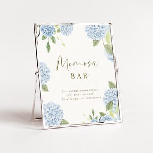May include: A white sign with blue hydrangea flowers and green leaves. The sign says "Momosa Bar" and includes instructions for making a mimosa: "Pour yourself some bubbly, add some juice and top with fruit of your choice."