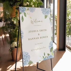 May include: A white wedding welcome sign with blue hydrangea floral accents. The sign reads "Welcome to the wedding of Joshua Smith & Hannah Bowen 16.10.2022 We're so glad you're here."