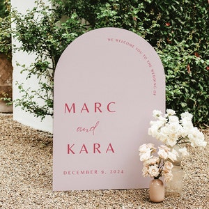 Könnte beinhalten: Ein rosafarbenes, bogenförmiges Hochzeitsschild mit dem Text "We welcome you to the wedding of Marc and Kara December 9, 2024" in weißen Buchstaben.