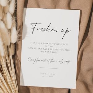 May include: A white rectangular sign with black text that reads "Freshen up" and other text below. The sign is on a brown fabric surface, with dried flowers on the left side.