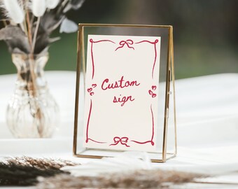 Pode incluir: Um letreiro de vidro com moldura dourada, fundo branco e escrita cursiva vermelha que diz "Custom sign". O letreiro é decorado com laços e volutas vermelhas.