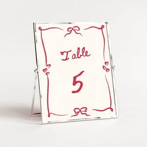 Puede incluir: Un porta números de mesa plateado con fondo blanco y letras rojas. El porta números tiene un marco decorativo con un lazo en la parte superior y las palabras "Mesa 5" escritas en el interior.