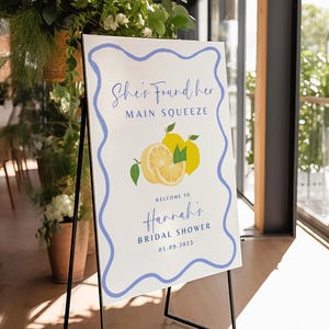 Puede incluir: Un letrero blanco con un borde ondulado azul sobre un caballete negro. El letrero dice "She's Found Her Main Squeeze" encima de una ilustración de limón. Debajo, dice "Welcome to Hannah's Bridal Shower 01.09.2023".