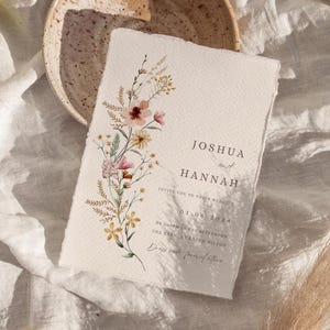 Può includere: Un invito di nozze color crema con un design floreale ad acquerello. L'invito presenta i nomi "Joshua e Hannah" e i dettagli del matrimonio. L'invito è posto su una superficie di lino bianco con una ciotola di ceramica e una fetta di arancia.