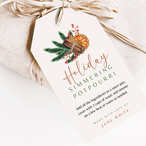 May include: A gift tag with the words "Holiday Simmering Potpourri" in red script. The tag features a watercolor illustration of cinnamon sticks, orange slices, pine needles, and red berries. The tag is tied to a wrapped gift with raffia.