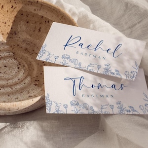 May include: Two white place cards with blue floral designs and names Rachel Eastman and Thomas Eastman in elegant script. The cards are on a white cloth, near a speckled ceramic bowl.