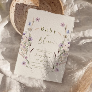 Pode incluir: Um convite para um chá de bebê com um design floral. O convite diz "Baby in Bloom" e inclui a data e hora do chá de bebê. O convite é impresso em papel texturizado.