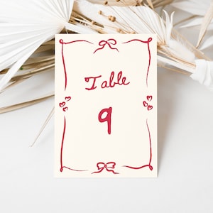 May include: A white card with a red border and red hearts. The card says "Table 9" in red cursive writing.