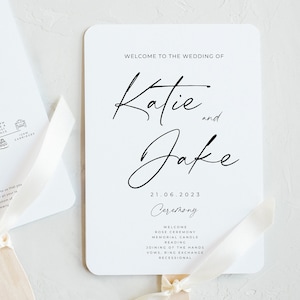May include: A white wedding program with rounded corners. The program features the names "Katie and Jake" in elegant script, along with the date "21.06.2023" and a ceremony schedule. A cream ribbon is attached.