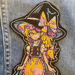May include: Embroidered patch showing a colourful anime-style witch character. The patch has a black border and depicts a girl in a witch hat, dress, and apron, with pink, yellow, blue, and purple accents. The patch is affixed to a denim jacket.