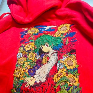 Può includere: Una felpa con cappuccio rossa con un ricamo in stile anime. L'opera d'arte raffigura un personaggio con i capelli verdi e un vestito rosso, circondato da girasoli e altri fiori. La felpa ha un cappuccio con coulisse.