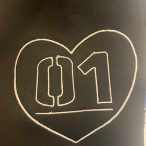 Puede incluir: Tela negra con un coraz&oacute;n bordado en blanco que contiene el n&uacute;mero "01" en una fuente estilizada.