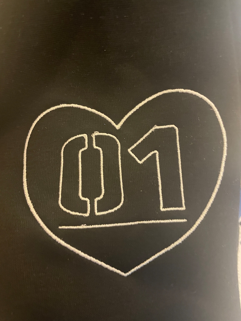 Puede incluir: Tela negra con un coraz&oacute;n bordado en blanco que contiene el n&uacute;mero "01" en una fuente estilizada.